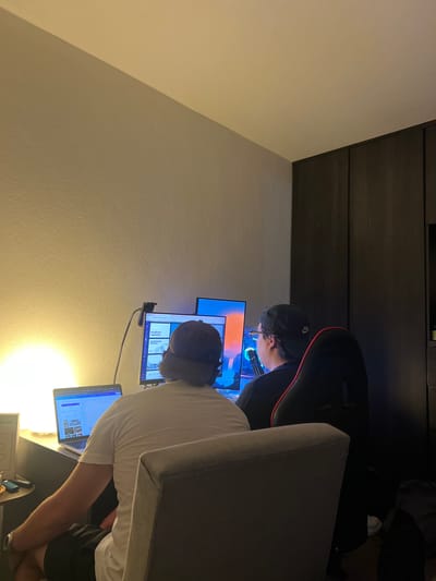 The image shows two individuals in an indoor setting, possibly a hotel room or workspace. One person, wearing a black cap, sits in a gaming chair with a red trim, focused on a multi-monitor setup with blue lighting emanating from the equipment. A second individual, wearing a white shirt and a cap, sits at a desk with a laptop and several screens. The location is presumed to be in Cholula, Mexico, though this can't be definitively confirmed from the image alone. The room features neutral-colored walls and dark wood paneling. There are cables and electronic devices scattered on the desk. The overall impression is of a workstation environment, with a focus on technology and screen displays.