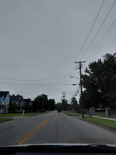 The image depicts a street scene in Lawrenceburg, United States of America. The sky is overcast and gray. The road, with a broken yellow line down the center, appears to be a two-lane street. On the left side of the road, several houses with pitched roofs are visible, some with American flags displayed. Trees line the street and the front yards of the houses. On the right side of the road, a sidewalk runs alongside a grassy area with stone pillars and a fire hydrant. A telephone pole with numerous wires stretches across the upper portion of the image, with another telephone pole visible further down the street. A few cars are present, one in the distance on the road, and the perspective suggests the photo is taken from inside a vehicle. The overall impression is of a quiet residential street on a cloudy day.