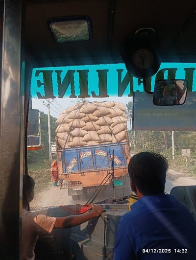 The image depicts a daytime scene viewed from the interior of a vehicle, likely a bus, on a rural or semi-rural road in Kaliganj, Bangladesh, dated 04/12/2025 at 14:32.
In the immediate foreground, within the vehicle from which the photograph was taken, a partial view of the dashboard and steering wheel is visible. A male individual in a light-colored shirt is positioned to the left, with his hands on the steering wheel, indicating he is the driver. Another male individual, wearing a blue shirt, is seated to the right, observed from behind, facing forward. The vehicle's front windshield has a blue tint, reflecting the mirrored text "ENIL LINE" at the top. An overhead light fixture and a small clock are mounted on the ceiling. A rearview mirror hangs to the right, reflecting the face of a person, likely a passenger or the blue-shirted individual.
Directly ahead, in the midground, an overloaded, rust-colored truck with a blue cargo bed is traveling on an unpaved or dusty road, moving in the same direction as the viewing vehicle. The truck's cargo bed is heavily stacked with numerous large, light-brown burlap sacks, secured with ropes, creating a significantly tall load. The rear of the truck displays yellow Bengali script and decorative patterns on the tailgate. Dust plumes are visible around the truck's wheels, consistent with movement on a dry, unpaved surface.
To the immediate left of the overloaded truck, near the edge of the road, a person wearing a patterned red and green garment stands observing the scene. The road surface appears uneven and unpaved, transitioning into light vegetation and sparse trees lining the roadside in the background. The sky is clear and bright.