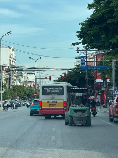 The image captures a street scene in Hoa Lư Ward, Vietnam, under an overcast sky. A red and yellow bus, labeled "NHO QUAN - ME - NINH BINH, VIEN 700 GIUONG" and with license plate "35F - 001.51," is prominent in the foreground, stopped at what appears to be a traffic signal. To its right, a green forklift is also stopped, with two individuals visible inside.
A turquoise car is positioned behind the bus, and numerous other vehicles, including cars and motorcycles, are present on the multi-lane road. Several traffic lights are visible, with at least one displaying red. Overhead, power lines crisscross the sky, and streetlights are mounted on poles. Buildings line the street, some appearing to be hotels or commercial establishments, with visible signage including "VINAPHONE." Trees and foliage are present, contributing to the urban landscape. People are seen on motorcycles and walking, suggesting normal daily activity. The road surface is asphalt.