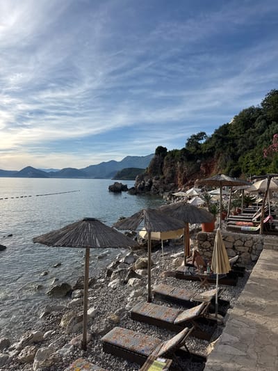 The image depicts a tranquil beach scene in Drobnići, Montenegro, under a partly cloudy sky with wispy cirrus clouds. The calm, azure sea gently laps against a rocky shoreline lined with straw umbrellas and lounge chairs. In the foreground, several lounge chairs with colorful patterned cushions are arranged on the pebbled beach, some shaded by rustic umbrellas. A person is seated on a lounge chair further back, appearing relaxed. Beyond the beach, a rocky cliff face covered in lush green foliage rises steeply. In the distance, layers of mountains create a picturesque backdrop. The overall atmosphere is one of peaceful relaxation and natural beauty.