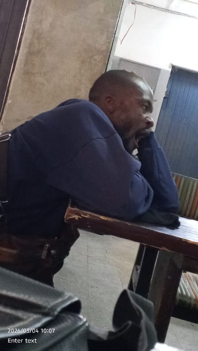An interior image depicts a male individual seated at a wooden table in Dakata, Nigeria. The man, with dark skin, short hair, and a beard, wears a dark blue long-sleeved garment. He is positioned facing right, with his right elbow resting on the table and his right hand supporting his chin/jaw area, his mouth slightly open. Behind him, to the right, a chair with vertical green, orange, and white stripes is partially visible. The table exhibits wear along its front edge. The floor is composed of light-colored tiles. The background on the left features a textured, light beige wall. To the right, a white wall is visible with exposed red wiring near the top, an electrical box, and a section of a dark blue corrugated surface. A dark, out-of-focus object is present in the immediate foreground at the bottom left. The image bears a timestamp of "2026/03/04 10:07".