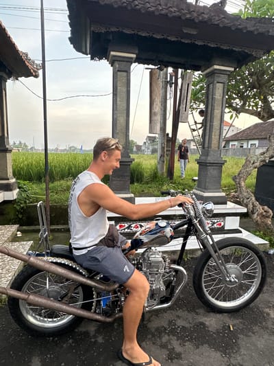 An outdoor scene in Celuk, Indonesia, depicts a blond-haired male seated on a custom motorcycle on a paved surface. He wears a white sleeveless top, gray shorts, dark flip-flops, and a dark sling bag across his chest, smiling towards the left. The motorcycle features a black frame, chrome engine and fork springs, a fuel tank with an eagle-themed design, a ribbed brown seat with a chrome sissy bar, and dual rusty exhaust pipes. In the mid-ground, behind the motorcycle, lush green rice paddies extend towards the horizon under an overcast sky. Traditional Balinese stone structures with dark, carved roofs are visible on both sides of the image. A utility pole stands centrally. To the right of the utility pole, a person on a metal A-frame ladder appears to be working near a stone pillar. Further back and to the right, another individual stands in the grass, smiling towards the viewer. Houses with tiled roofs are visible in the distance. A gnarled tree trunk is positioned on the right. A small, white object resembling a cigarette butt lies on the pavement near the motorcycle's front wheel.