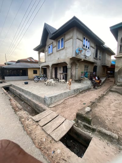 This image captures a street scene in Enwan, Nigeria, under an overcast sky. A prominent two-story concrete building with a dark grey pitched roof and blue-tinted windows dominates the center-right. Two "SPACE" branded satellite dishes, one light blue and one white, are mounted on its upper facade. The ground floor features patterned concrete tiling near an entrance. To the left, a single-story building with a corrugated metal roof and a black-and-white checkered wall section is visible.
In the foreground, a concrete drainage ditch, partially filled with debris and stagnant water, runs parallel to the viewer. Three wooden planks form a makeshift bridge over a wider section of this ditch, and a rectangular opening into a deeper cavity is visible further right. The ground is unpaved, consisting of reddish-brown dirt with scattered litter.
A raised concrete patio occupies the area in front of the main building. On this patio, a yellow and black motorcycle is parked. Several goats are present: two white adult goats and a white kid are near the motorcycle, and a light brown and white goat stands on a raised concrete ledge to the right. A dark goat is also visible further back near the motorcycle.
Four individuals, appearing to be men and children, are seated casually on a concrete ledge or bench to the right of the main building. Clothes are draped on a railing or line near them. Overhead power lines and a utility pole are visible against the sky on the far left. A black plastic basin sits on the concrete patio near the motorcycle. A single green flip-flop lies on the dirt path.