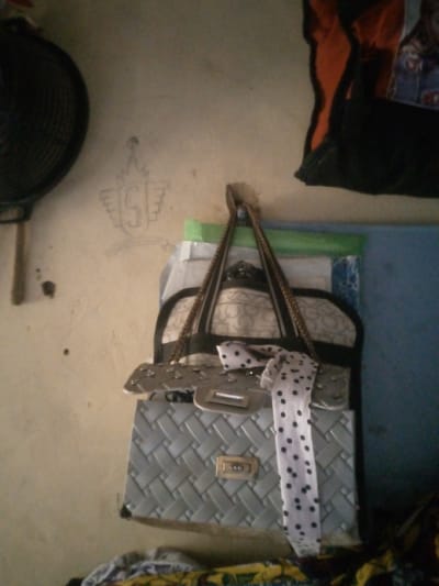The image depicts a dimly lit indoor scene, likely a bedroom or storage area in Unguwan Rimi, Nigeria. A grey woven handbag with a gold clasp is the central object, hanging from a braided strap. A white and black polka dot scarf is draped over the handbag. To the left of the handbag, a portion of a black fan is visible, and a stylized drawing of what appears to be the letter "S" within a crown-like emblem is on the beige wall behind it. The background includes other indistinct objects, including a blue wall section and some fabric. The lighting is poor, creating shadows and obscuring finer details. There are no people visible in the frame.