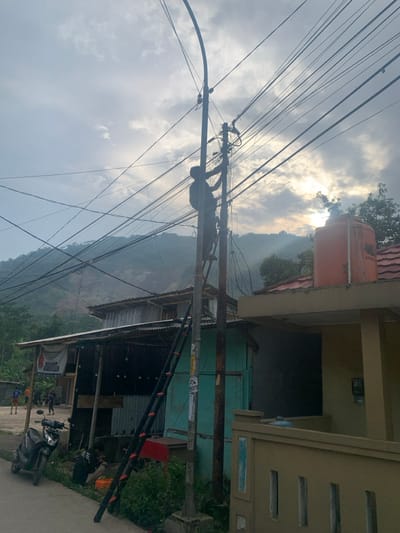 The image shows a man working on a utility pole in the city of Lagadar, Indonesia, at coordinates -6.924 latitude and 107.537 longitude. He is standing on a ladder and appears to be connecting wires. The pole is located between two buildings. The building on the left is partially obscured but appears to be a shop. The building on the right is a house. The scene is situated on a road with a motorcycle parked in front of the shop. In the distance, a mountain range can be seen. The sky is overcast, and the sun is shining through the clouds. There are many power lines running across the sky. The image was taken in the afternoon.