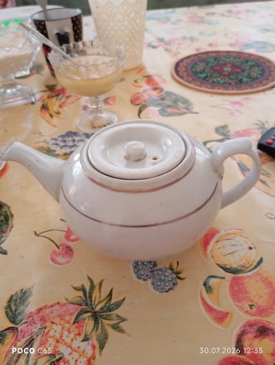 The image shows a white teapot with a gold trim on a tablecloth with a fruit pattern. The teapot is in the center of the frame. In the background, there are two glass dishes, one containing a light-colored liquid and the other containing a white granular substance, and a mug. There is also a decorative coaster on the tablecloth to the right of the teapot. The scene appears to be set indoors, likely on a table. No people are visible in the image. The date and time stamp "30.07.2026 12:35" is present in the bottom right corner, along with "POCO C65" in the bottom left. The location of Beruniy, Uzbekistan, is not indicated in the image itself.