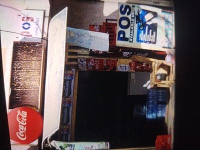The scene depicts an outdoor commercial vending stall or shop frontage situated in Kaura Goje, Nigeria. The establishment offers Point-of-Sale (POS) services, evidenced by a prominent sign displaying "POS AVAILABLE HERE" and detailing services such as "CASH DEPOSIT," "WITHDRAWAL," "BILLS PAYMENT," and "TRANSFER." Another "POS AVAILABLE" sign is partially visible on the upper left.
The stall's merchandise includes stacked red Coca-Cola crates, some containing bottles, positioned on the right side. Several blue plastic crates filled with bottled water are visible below these crates. A red circular sign featuring the Coca-Cola logo rests on a wooden surface to the far left. Display units feature "Hollandia" branding. A dark, rectangular opening, likely a counter or service window, is centrally located, with a gray refrigerator or cooler visible behind it to the right. Various smaller packaged goods, including an item branded "Sweet Me" with "0000" below it, are arranged around the stall. A large, reflective, silver-colored object, possibly a metal sheet, is prominent on the left side of the frame, partially obscuring other elements. No individuals are present in the image, and no specific actions or interactions are depicted.