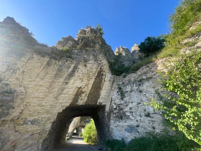 The media file captures a view of a road passing through rock formations, likely a natural or man-made tunnel, in Asparuhovo, Bulgaria. The scene is dominated by rough, light-colored stone walls that appear layered and textured. Sunlight streams in from the right side of the frame, creating a bright glare on the rock face and casting shadows within the tunnel opening.
The road is paved and curves into the distance, passing through at least two distinct archways or tunnels carved into the rock. Lush green vegetation, including trees and bushes, grows on the slopes and around the entrances of the tunnels, indicating a natural setting with a path or road created within it. The sky above is a clear, vibrant blue, suggesting fair weather and a daytime setting, likely late morning or early afternoon due to the angle of the sunlight.
There are visible markings on the rock walls, appearing to be graffiti. Some inscriptions are in Cyrillic script, though they are not entirely legible. For example, on the left wall, some faint markings can be seen, including what might be "ВАРНА" and "6 ЕВГЕНИ". Other scribbled graffiti, some in blue and red, is also present on the rock faces.
The overall impression is one of a scenic and somewhat rugged natural landscape where human passage has been facilitated by carving through the rock. The image conveys a sense of exploration and the beauty of natural formations. No people or specific events are depicted, focusing solely on the environment.