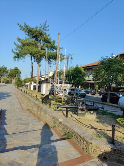 A sunny daytime scene unfolds in Litochoro, Greece, along a paved walkway lined with stone and a chain fence. In the foreground, a shadow of a person is cast on the stone steps leading down. To the right of the walkway, an old ship's anchor and a partial skeletal replica of a boat are displayed, adorned with lights. Beyond the display, a street runs parallel to the walkway, with parked cars and a motorcycle. Buildings with brown-tiled roofs and balconies line the street, interspersed with lush green trees. The sky is a clear, bright blue, indicating fair weather. The overall atmosphere is peaceful and scenic, suggesting a public area or promenade in a coastal town.