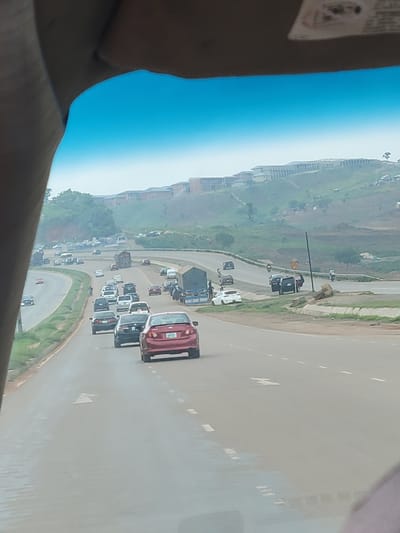 This image depicts a highway scene in Kugbo, Nigeria, characterized by moderate traffic congestion. The road is a multi-lane highway, with vehicles traveling in various directions. In the foreground, several cars are visible, including a red sedan in the right lane, followed by a black sedan and another dark-colored car. Other vehicles, including trucks and cars, are dispersed throughout the roadway.
The environment appears to be a hilly area, with buildings constructed on the slopes in the background. Vegetation is present on the hillsides and along the sides of the highway. The sky is visible at the top of the image, appearing overcast or hazy. The overall impression is that of a busy urban or suburban road network in Nigeria.