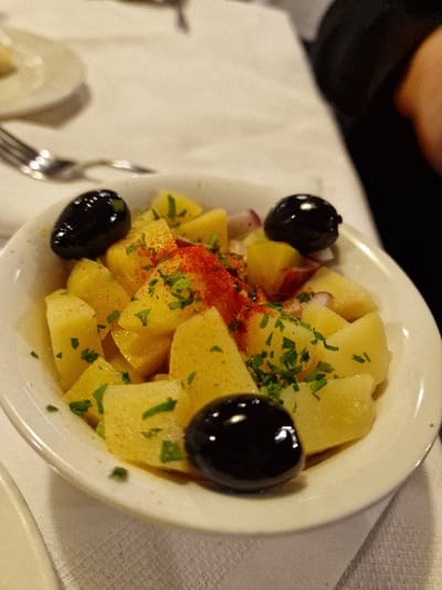 The image captures a close-up view of a potato salad served in a white, circular bowl. The salad consists of yellow, cubed potatoes, garnished with chopped green herbs, consistent with parsley, and a central sprinkling of red paprika. Three whole, glossy black olives are positioned on the surface of the salad. Interspersed with the potatoes are small pieces of reddish-purple material, likely red onion. The bowl rests on a white, textured tablecloth. To the upper left, a silver fork and a partially visible white plate are present. A dark-colored garment, likely a sleeve, is visible on the right side of the frame, indicating a person seated at the table. The scene depicts a dining setting, consistent with a restaurant in Odorheiu Secuiesc, Romania.