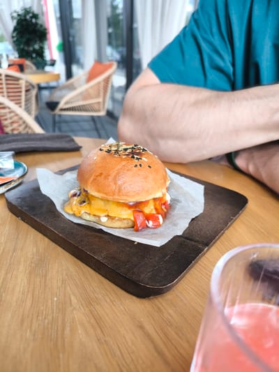 A close-up shot shows a burger served on a wooden board on a wooden table. The burger features a sesame seed bun, cheese, and red sauce. A glass with a pink beverage is also on the table, partially visible in the lower right corner. An individual wearing a teal shirt is partially visible in the background. The background includes furniture, potentially outdoor seating, and a glimpse of trees through a window. The location is Fryazino, Russia.