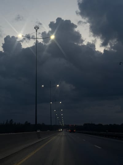 The image depicts a road in Kondratovo, Russia, at dusk or night. A multi-lane highway stretches into the distance, bordered by a concrete barrier on the left and trees or vegetation on the right. Streetlights, some with a double-headed eagle emblem on the tallest pole, illuminate the road. The sky is dominated by dark, heavy clouds, with hints of lighter sky visible through breaks. A few vehicles are present on the road, indicated by their headlights and taillights. The overall atmosphere is moody and dark, suggesting a potentially overcast or stormy evening.