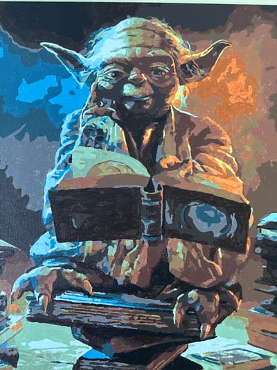 The image is a painting of Yoda, a fictional character from Star Wars, sitting and reading a book. He is depicted in the style of a paint-by-numbers artwork, with distinct blocks of color. Yoda's face is a mixture of greens and browns, with large ears characteristic of the character. He is wearing a brown robe. His hands are holding open a dark-colored book, which has a circular blue design on one of its pages. The background is abstract, with areas of blue on the left and orange and brown tones on the right, suggesting a stylized environment. There are also stacks of what appear to be books or similar objects in the foreground and to the right of the frame. The painting has a textured appearance, consistent with acrylic paint on canvas. There is no indication of the city of Tambov, Russia, or any specific urban or geographical location within the image.