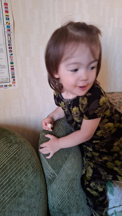 A young girl with brown hair and fair skin is leaning on a green upholstered sofa. She is wearing a dark dress with a green floral pattern. Her right hand is holding a small piece of food, and her mouth is slightly open. The background features a textured wallpaper and a poster on the left wall with flags of various countries. The image appears to be taken indoors in a residential setting. The poster on the wall is part of an educational display related to geography or international affairs, potentially in a home or classroom environment in Mednoe-Vlasovo, Russia.