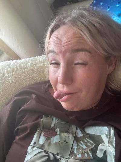 A young woman with blonde hair is lying down on a fluffy white cushion, possibly on a couch or bed. She is wearing a brown t-shirt with a printed image of clothing and shopping bags. The woman is winking and sticking her tongue out playfully. The background is blurred but appears to be an indoor setting. There is no indication of the location being Pozdyshki, Russia.