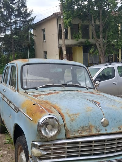 The image shows the rusted front of an old, blue car with a prominent chrome grill. "BUCKINGHAM" is vertically spelled out on the vehicle next to the grill. The windshield is cracked. The car appears to be parked on a street in the city of Baylovo, Bulgaria, as indicated by the architecture of the adjacent buildings and the presence of trees in the background. Another modern, silver car is parked nearby. There are no people visible in the immediate frame. Power lines run through the image as well.