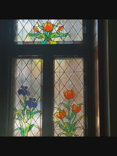 The image shows a close-up of a stained glass window with a floral design. The window is divided into several panes, most of which are filled with a diamond-patterned clear glass.
The top pane features a stained glass motif of orange tulips with green leaves and stems, arranged in a decorative manner.
The two lower panes depict more stained glass floral designs. The left pane contains blue irises with green leaves and stems. The right pane features orange tulips with green leaves and stems. The floral elements are rendered in vibrant colors against the textured clear glass. The entire stained glass panel is set within a dark wooden frame. The lighting suggests the window is illuminated from behind, casting a warm glow on the colored glass.
The image does not provide any visual information to determine its location in the city of Saint-Août, France.