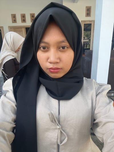 A medium shot captures two young Indonesian women in hijabs, standing outside what appears to be a building in Jambearum, Indonesia. The woman in the foreground, positioned centrally, is wearing a black hijab and a light-colored jacket. Her gaze is directed at the camera, and she has a neutral expression. Behind her, to the left, a second woman, also in a hijab (this one white), is partially visible, facing away from the camera and looking down. The background reveals the exterior of a building with wooden window frames and decorative elements. The lighting suggests it is daytime, casting subtle shadows.