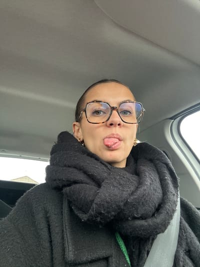 A female individual is depicted from the chest up, seated inside a vehicle in the city of Saint-Cyprien, France. She has her hair pulled back and is wearing large, square-framed eyeglasses with a tortoise-shell pattern over light-colored eyes. Her tongue is extended from her mouth. She is dressed in a dark, textured coat and a large, dark, knitted scarf wrapped around her neck and shoulders. A grey vehicle seatbelt is positioned diagonally across her right shoulder and chest. Gold-colored earrings are visible on both ears. A bright green strap or cord is partially visible beneath her coat on the left side. The vehicle's interior roof lining is light grey, and a blurred exterior view through a window on the left shows a light-colored structure beneath an overcast sky.