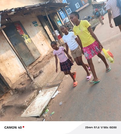 The image portrays three young females walking together on a paved street in Agbani, Nigeria, under the soft, warm light of what appears to be late afternoon or early morning. The scene captures an everyday moment in a bustling street environment.
From left to right, the youngest girl wears a pink t-shirt with "AMAZING..." partially visible text, a plaid skirt, and bright orange slides. Beside her, a slightly older girl sports a white t-shirt, khaki shorts, and pink slides. The eldest girl, on the right, is dressed in a yellow t-shirt and a pink floral-patterned skirt, carrying a translucent yellow plastic bag, and wearing green flip-flops. The girls' expressions are neutral, indicating a casual pace or daily activity. Other blurred figures and market-like structures are visible in the background, suggesting a lively community setting.
The street is flanked by rustic, somewhat dilapidated buildings with corrugated iron roofs and weathered walls. Some structures feature large, closed metal doors, possibly storefronts, with faded posters or graffiti. Scattered debris, including cardboard and wrappers, lies on the ground near the buildings and on the street. The general architectural style and street conditions are consistent with an urban or semi-urban setting in Nigeria. The light clothing and open footwear of the subjects suggest warm weather.
Text at the bottom left corner indicates "CAMON 40," likely the camera model used. At the bottom right, photographic metadata "23mm f/1.8 1/180s ISO50" is displayed.