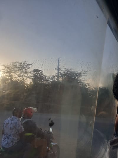 Two people on a motorbike are captured on a sunny day, likely near trees and a utility pole. A man in a floral shirt stands beside the motorbike, while the driver wears a helmet and black clothing. The image is taken through a transparent surface, creating distortions and reflections. The lighting is bright, casting shadows and highlighting the subjects.