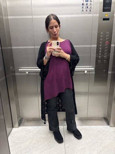 The image shows a woman taking a selfie in an elevator. The elevator has stainless steel walls and a panel of buttons numbered 0 through 10. The button for floor 1 is illuminated. The woman is wearing a purple top, black pants, and black shoes. She also has a black shawl or cardigan and several bracelets on her left wrist. She is holding a light-colored smartphone with decorative elements on the back. The floor of the elevator appears to be light-colored marble. The location in San Isidro, Peru cannot be definitively determined from the image.