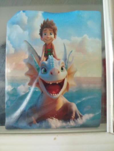 The image shows a digital illustration featuring a young boy with brown, spiky hair and a wide smile, wearing a green and red outfit. He is riding on the back of a friendly-looking, light blue dragon with green eyes and large, open jaws. The dragon appears to be in water, with waves and spray around it. The background depicts a bright blue sky with fluffy white clouds and a hint of a distant horizon, suggesting a daytime outdoor scene. The illustration is framed by a white border, and the overall impression is one of a cheerful and adventurous cartoon. There is no visible indication of the location being in Крывасельскі сельскі Савет, Belarus, as the scene is an animated illustration and not a real-world photograph.