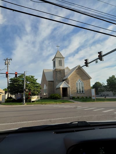 This is a rotated image of a church and a street scene, taken from a vehicle. The primary subject is a stone church with a steeple and a white roof. A sign in front of the church reads "Sunshine United Methodist Church" and displays service times. The church is situated next to a paved road with white lane markings.

Several traffic lights are visible, with some indicating red. Overhead power lines run across the top of the frame, creating a dark, linear pattern against a blue sky with scattered white clouds. Trees and green grass are present, suggesting an outdoor setting. The image appears to be taken during the daytime, likely late afternoon or early evening given the lighting. No people are clearly visible.

Text on a sign reads "Berea United Methodist CHURCH SUNDAY WORSHIP 9 AM-8 PM". Another sign is partially visible and has "SUNSHINE" on it. A traffic signal sign shows the number "6". The overall scene suggests a typical town street with a church and traffic infrastructure. The rotation of the image makes it difficult to ascertain the exact orientation of the scene.