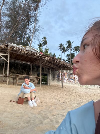 The media file shows a selfie of a person with their lips puckered as if blowing a kiss, with their face taking up the top left portion of the frame. In the background, a sandy beach is visible with palm trees and a rustic, open-air hut with a thatched roof. The hut appears to be made of wood and bamboo.
Further back, there are hints of other structures and possibly people gathered, suggesting a public area. The sand shows footprints, indicating recent activity. In the mid-ground of the image, a second person is sitting on the sand, looking down at their phone, and holding a brown purse. This person is wearing a light blue collared shirt and light-colored shorts, with white sneakers.
The weather appears overcast with a cloudy sky. The presence of palm trees and the hut suggests a tropical or subtropical coastal location. The overall mood is relaxed and casual, capturing a moment of leisure. There are some graffiti markings visible on a wall behind the hut, but the text is difficult to decipher clearly. The time of day appears to be daytime, given the lighting conditions. The image is oriented sideways.