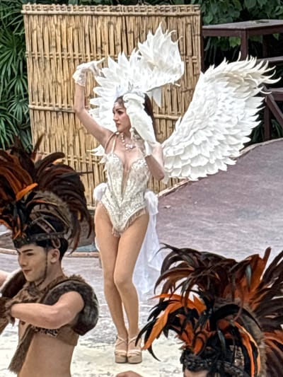 In Nongprue, Thailand, a theatrical performance is underway on an outdoor stage. The central figure is a performer dressed as an angel, adorned with large white feathered wings and a matching headdress. They wear a shimmering, form-fitting costume with intricate beadwork and a jeweled necklace. The performer is in mid-pose, with one arm raised and gloved, their body tilted slightly to the side.
Flanking the central performer are other individuals in elaborate costumes. To the left, a male performer is visible from the chest up, wearing a headdress of large, dark feathers with touches of orange and brown. They have fur or fur-like material draped over their shoulders and chest. To the right, only the top of another performer's head is visible, also wearing a feathered headdress with dark and orange plumes.
The stage backdrop consists of a woven bamboo screen, and behind that, lush green foliage and a wooden structure are visible. The stage floor appears to be a light-colored, textured surface. The lighting suggests it is daytime, with natural light illuminating the scene. The overall atmosphere is one of vibrant performance and cultural display.