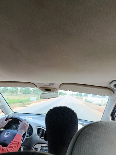 The image captures the interior of a Volkswagen car driving on a road in Kafuri, Nigeria. The perspective is from the back seat, looking forward. A person with dark, curly hair is in the driver's seat, wearing an orange shirt and gripping the steering wheel. Their hands are visible on the wheel, and the dashboard displays various gauges. The road ahead appears to be paved and leads through a rural landscape with trees and some buildings in the distance. The sun visors are down on both sides. The overall scene depicts a typical journey within the town.