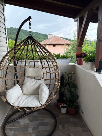 This image captures a serene outdoor living space, likely a balcony or porch, overlooking a picturesque landscape in Polena, Bulgaria. The central subject is a comfortable hanging egg chair, featuring a dark metal frame intricately woven with natural-colored rope. It is adorned with several plump, white cushions and pillows, inviting relaxation.
The chair rests on a grey-toned, patterned tiled floor. To the right, an assortment of potted plants adds greenery to the space; these include various types of foliage plants in terracotta and white pots, positioned on the floor and a low ledge. Further along the balcony edge, a white planter box contains vibrant red flowering plants. The balcony area is partially sheltered by a wooden roof structure with visible beams and supports.
Beyond the immediate outdoor space, the background reveals a tranquil, verdant landscape. Rolling green hills or mountains, densely covered with trees, dominate the view under an overcast sky with soft clouds, suggesting a mild daytime. Nestled within this natural setting, glimpses of buildings with distinctive reddish-orange tiled roofs are visible, indicative of a rural village or town. No people or visible text are present in the image. The scene evokes a sense of peaceful enjoyment of nature and comfortable outdoor living.