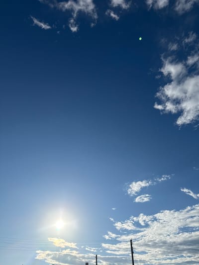 This media file is an outdoor photograph taken on a clear, sunny day in Уральское, Russia. The primary subject is a bright blue sky with scattered white clouds. The sun is a prominent feature, shining intensely and creating a glare in the lower right portion of the image. A thin band of clouds, elongated and streaky, are visible on the right side, some appearing backlit by the sun.
In the upper left quadrant of the image, there is a distinct, small, bright green circular object. Its nature is unclear from the image alone, but it stands out against the deep blue sky. Towards the right side of the image, there are visible signs of human infrastructure. A utility pole with multiple wires running across it is present, suggesting the location is near a populated area or along a transportation route.
The time of day appears to be daytime, likely late morning or early afternoon, given the position of the sun and the bright lighting conditions. The weather is fair and sunny. There are no people or animals clearly visible in the image, and no text is present. The overall impression is one of a serene, expansive sky with a touch of mystery introduced by the green object.