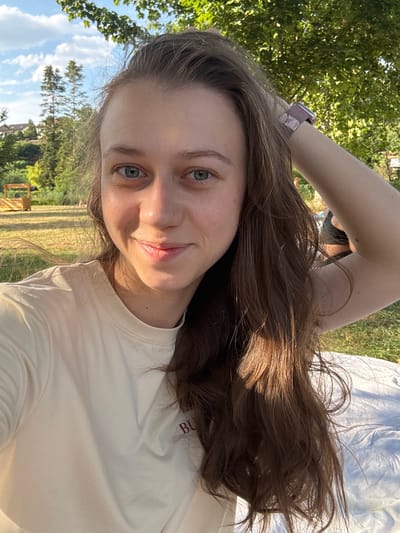 A young woman with long, brown hair is lying on her back outdoors. She is wearing a light-colored t-shirt. Her right arm is raised behind her head, and she is holding her phone for a selfie. The background shows trees, grass, and a blue sky with clouds. The sun is shining, casting shadows and highlighting her hair. The setting appears to be a park or grassy area. On her left wrist, she is wearing a pink smartwatch. A red embroidered logo with the letters "BU" is visible on her t-shirt. The image has a casual and relaxed feel, suggesting a sunny day activity outdoors. The location is identified as Bürgstadt, Germany.
