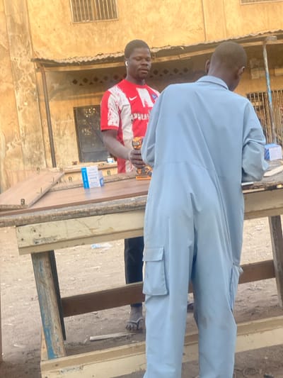 Two men stand outdoors near a workbench. One man wears a light blue jumpsuit and faces away from the camera. The other wears a red and white athletic shirt and holds a drill. The workbench is surrounded by wooden planks. Boxes are placed on the bench. A building with a window and metal grate is in the background. The ground is unpaved.
