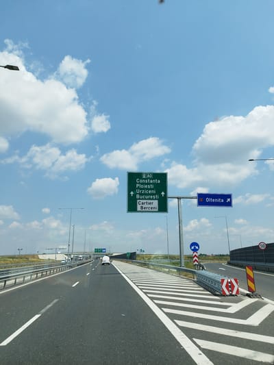 The image shows a highway in Popești-Leordeni, Romania. A green highway sign indicates directions to Constanța, Ploiești, Urziceni, București, and Cartier Bêrceni. A blue sign points towards Oltenița. The highway is paved and has white lane markings. There are guardrails along the side of the road, and several vehicles are visible on the highway, including a white van. The sky is blue with white clouds. There are streetlights and traffic signs visible along the road. A speed limit sign of 50 is also visible. The scene appears to be a sunny day.