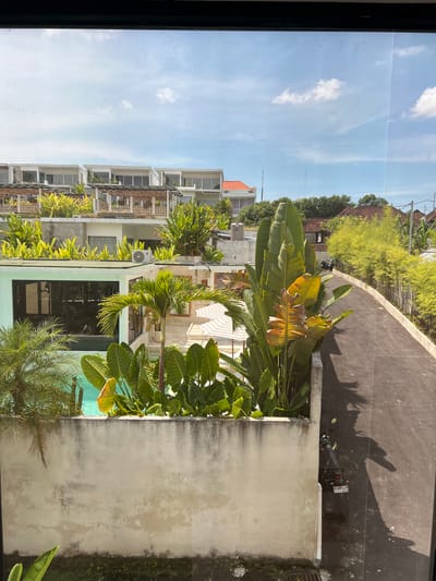 The image shows an outdoor scene in Buduk, Indonesia, as viewed from a high vantage point, possibly a window. The sky is visible with light clouds. Buildings with multiple balconies and greenery on each level are seen to the left. Lush tropical plants are prominent in the midground, including banana trees and palm trees, partially obscuring what seems to be a swimming pool. A gray concrete wall is on the right. Further up, a paved road, likely for vehicles, runs parallel to the scene, and a parked motorbike is visible on the road. Above the road, there are additional buildings obscured by more dense tropical vegetation.