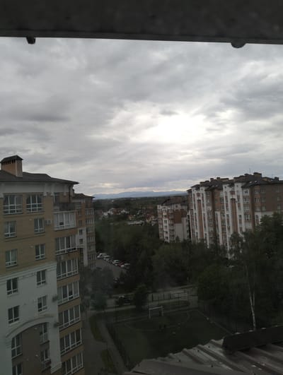 The image captures an elevated view from what appears to be an upper story of a building, overlooking a residential area in Krykhivtsi, Ukraine. The sky is overcast with a thick layer of grey clouds. Two multi-story apartment buildings dominate the left and right sides of the frame, each with a distinctive architectural style featuring beige and white facades and dark roofs. Between the buildings is a view of a town or village, with several smaller buildings that appear to be houses with red/brown rooftops surrounded by trees and a landscape showing hills in the distance. In the foreground, a soccer field with goals at each end is visible. Several trees partially obscure the view of the soccer field. A few people are visible on the field. In the background, a road is lined with parked cars, including what looks like an SUV.