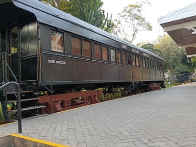 The image features an old train car, painted black with red undercarriage, prominently displayed on a brick-paved area, likely as a museum exhibit. The train car has the words "COCHE COMEDOR" on its side and "LIMA, MIRAFLORES, CHORRILLOS" along the top. A set of stairs leads up to a door on the left side of the car. The background contains lush greenery, trees, and a partial view of a building with a white roof. The presence of "MIRAFLORES" on the train car indicates that the location is in Miraflores, Peru. The setting suggests a park or historical site where the train car is preserved as a piece of local heritage.