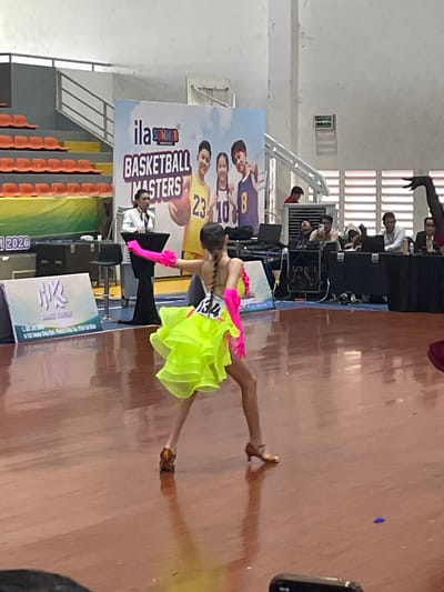 In the city of Thuận An, Vietnam, within what appears to be an indoor sports arena, a young female dancer is captured mid-performance. She is wearing a vibrant neon yellow costume with ruffled layers and bright pink arm coverings, and has the number "134" visible on her back. Her stance and the dynamic pose suggest she is engaged in a competitive dance event.
The background features a large banner with the text "ila SUMMER BASKETBALL MASTERS" and images of basketball players. Several other individuals are present in the background, including a woman in a white collared shirt standing behind a podium, likely an official or announcer. Other people are seated at tables with laptops and appear to be observing or working at the event. The flooring is a polished wooden surface, reflecting the lighting and the dancer. Orange stadium seating is visible in the upper left of the frame, indicating a spectator area. The overall environment suggests a formal event, possibly a competition or exhibition.