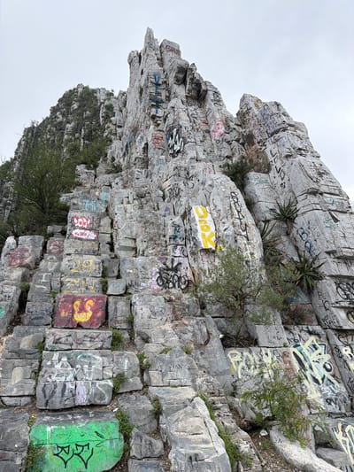 The image captures a massive, rugged cliff face, heavily covered in colorful graffiti, against an overcast, light gray sky. The cliff, composed of large, stacked gray rock formations, rises steeply, with various ledges and crevices.
Numerous patches of vibrant spray paint adorn nearly every visible rock surface. These include large, block-lettered words, intricate tags, abstract symbols, and some stylized drawings. Visible text and designs include a prominent yellow block with black letters that appear to spell "DIOX" or "DIOS." Further down, a reddish-orange block features yellow, stylized figures with round heads and simple features, while an adjacent bright green block displays two black, symmetrical designs resembling stylized faces or characters. Other visible text fragments and dates, such as "2021" and various illegible names or acronyms, are scattered throughout in colors like yellow, red, white, black, purple, and blue.
Sparse vegetation, including small bushes and agave-like plants, grows from the rock crevices and ledges, adding hints of green to the scene. The time of day is daytime, indicated by the bright, diffused light from the overcast sky. No people are visible. The scene depicts a natural rock formation in Santa Catarina, Mexico, transformed into a canvas by extensive human artistic expression.