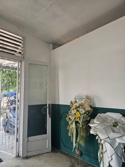 The image shows the interior of a building, likely a shop or restaurant, in Banyuurip, Indonesia. The room is sparsely furnished, with white walls and a dark green wainscoting. A white-painted door with a glass pane is open to the left, revealing a glimpse of the outdoors with foliage and a parked motorcycle. In the foreground, two flower arrangements stand on display. The one on the left is a bouquet of various flowers, including yellow, white, and peach-colored blooms, adorned with green ribbon and a white circular sign with green text that reads "Selamat & Sukses Atas Dibukanya Ayam Rempah Lek War". The arrangement on the right is a larger, more elaborate display wrapped in white paper with gold trim. The overall impression is one of a newly opened establishment, possibly a food business, as suggested by the flower arrangement's inscription. The lighting is natural, casting soft shadows. The floor is tiled in a light color with some dark staining. The ceiling shows some visible cracks and imperfections.