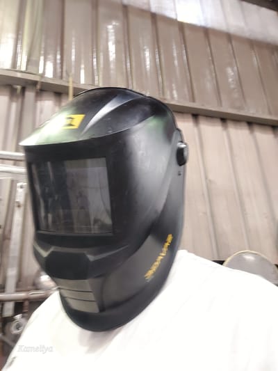A person wearing a black welding helmet is the central subject of this image. The helmet has a yellow logo on the top and a reflective visor. The person is wearing a white t-shirt. The background consists of a corrugated metal wall and some industrial shelving. The image is taken indoors, likely in a workshop or factory setting. The text "Kameliya" is visible in the bottom left corner of the image. The image's metadata indicates that it was taken in Alençon, France.