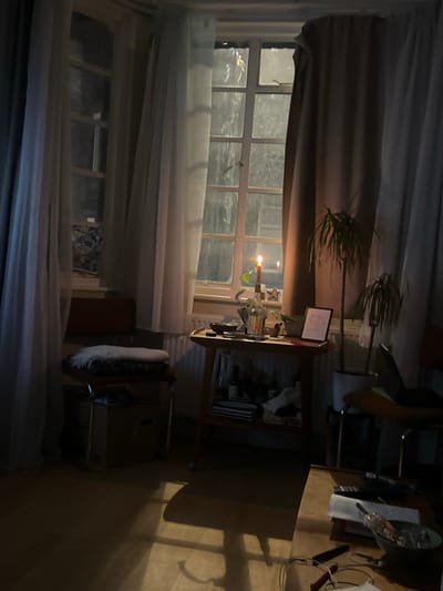 A dimly lit room features windows partially covered by curtains. Outside, trees are visible. An illuminated candle sits on a table next to the window. To the right of the table is a storage unit. A chair is situated beside a desk. Shadows stretch across the floor. The overall atmosphere is dark and subdued.
