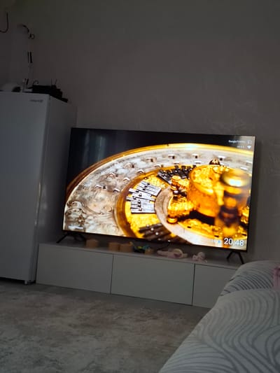 The image shows a television screen displaying a close-up of a golden, ornate object, possibly a mechanical music box or automaton. The object is intricately detailed with gears and decorative elements. The TV screen also displays information in the top left corner: "GoogleTV9995" and "Wi-Fi". In the top right corner, the time "20:48" and the temperature "19°" are visible. In the bottom right corner of the TV screen, the text "David F. Watson" is present, likely indicating the photographer or artist.
The television is placed on top of a white cabinet, which has a few small decorative items on it, including what appear to be plush slippers. To the left of the TV, there is a white refrigerator with the "ATLANT" logo visible on its side. Various cables and electrical items are also visible behind the refrigerator. The scene appears to be indoors, likely in a living room or entertainment area. The lighting suggests it is evening, given the displayed time. The overall setting is domestic, with a focus on the technological display of the television and the surrounding household objects.
