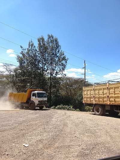 The image depicts an outdoor environment in Kiambu Township ward, Kenya, under a clear blue sky with scattered white cumulus clouds. The ground is a light brown gravel or dirt surface, with a small crumpled white object visible in the lower-left foreground. A white-cab, yellow-bed dump truck is positioned in the midground on the left, oriented towards the right, generating a prominent dust cloud around its rear wheels, indicating movement. To the right, the rear section of a second, stationary truck with a light-brown wooden cargo bed is visible. The background consists of dense green foliage, including tall trees and shrubs. Multiple horizontal power lines traverse the upper portion of the frame, and a utility pole with insulators is present in the upper-right quadrant. No people are visible.