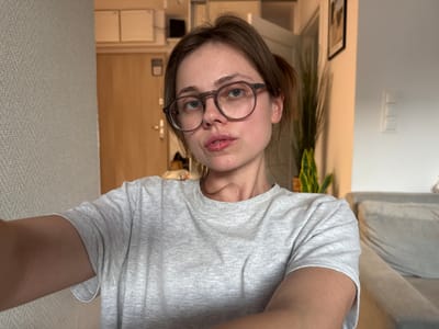 A young woman with brown hair pulled back and wearing glasses is taking a selfie in what appears to be an apartment. She is wearing a grey t-shirt. In the background, there is a doorway with a light-colored door, a textured wall, and part of a sofa. There is also a potted plant visible. The scene does not contain any elements suggesting a location in Szklarska Poręba, Poland.