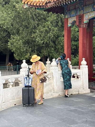 In Xicheng District, China, two women stand on a paved walkway in front of a white marble balustrade adorned with ornate carvings. Behind the balustrade, lush green trees form a dense backdrop. In the foreground, a woman on the left, wearing a yellow straw hat, a long yellow dress with pink floral patterns, and gold shoes, inspects a small purple fan in her hand. She has a necklace around her neck and a brown designer handbag with a gold chain strap slung over her shoulder. Next to her stands a black suitcase with a blue and white patterned bag on top, and a tripod attached to the suitcase with a small camera on top. To the right, a second woman, viewed from behind, wears a form-fitting dark green dress with a floral pattern in shades of teal and black. She has a blue hair accessory and black and white shoes. She leans against the balustrade, her right hand on her hair. A portion of a traditional Chinese pavilion with red pillars and a decorative, tiled roof is visible in the upper right corner of the frame. Further in the background, on the left, two other individuals are visible, one sitting and the other standing. The overall scene captures a moment of leisure and sightseeing in a culturally rich environment.